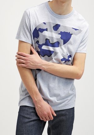 Homme portant un T-shirt gris clair avec un grand logo à motif camouflage bleu marine, jean foncé, debout les bras croisés sur la poitrine.