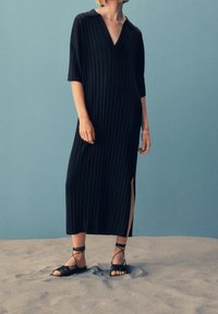 Black ribbed midi dress with a V-neck, short sleeves, and side slits. Worn with black strappy flat sandals. Background is a light blue.