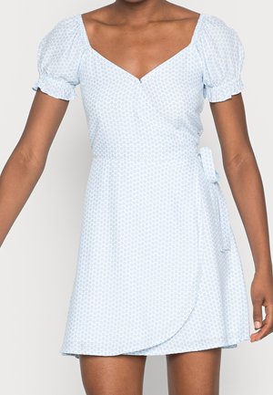 Nainen, joka pitää yllä vaaleansinistä, lyhythihaista kietaisumekkoa, jossa on pieni kukkakuosi ja V-pääntie, sidottu vyötäröltä oikealta puolelta.