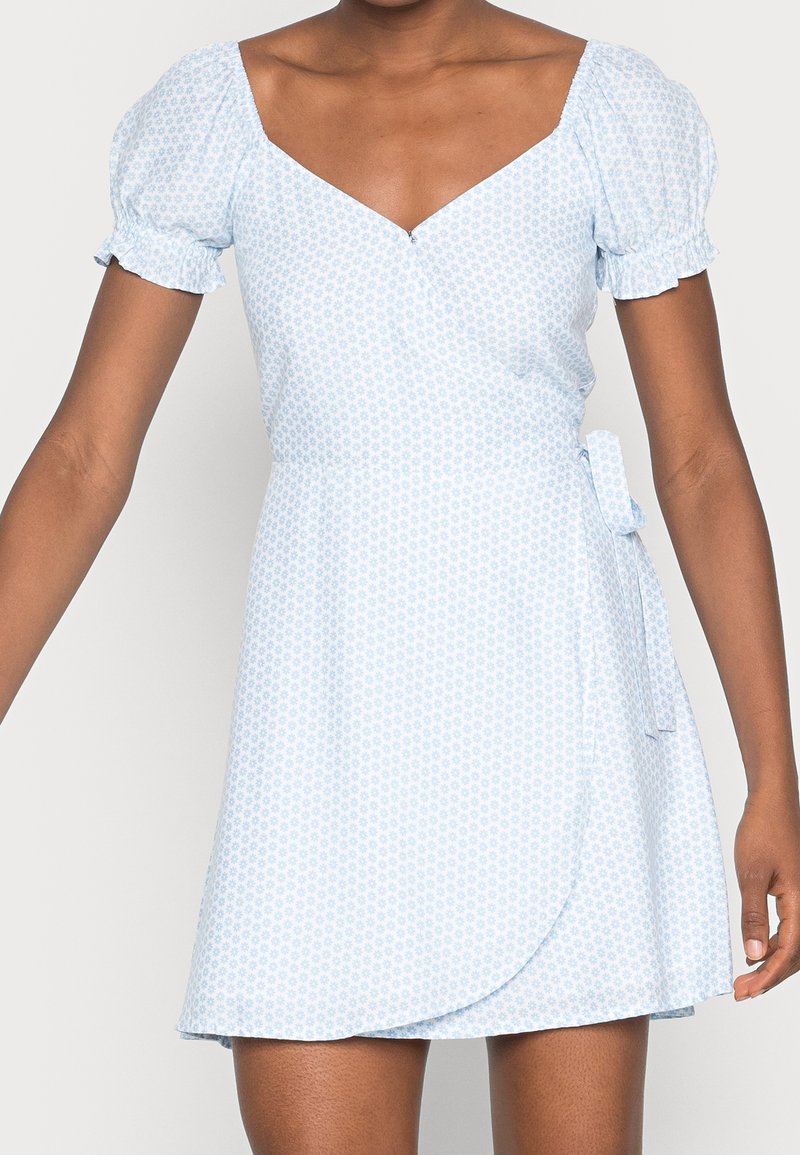 Femme portant une robe portefeuille bleu clair à manches courtes, imprimé floral petit, avec un col en V, nouée à la taille sur le côté droit.