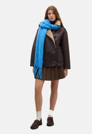 Bruine leren jas met beige kraag, blauwe sjaal, geruite rok, witte sokken en donkere schoenen. Zachte texturen en gelaagd ontwerp.