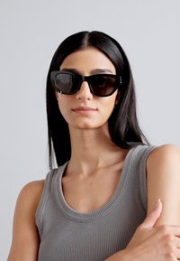 Occhiali da sole neri a forma di occhio di gatto con lenti scure, dotati di un design audace della montatura. Il modello indossa un top grigio a coste senza maniche.