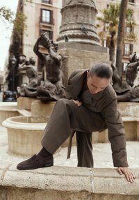 Frau in braunem Outfit klettert über den Rand eines steinernen Brunnens mit Skulpturen und Palmen in einem urbanen Platz.