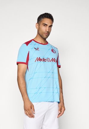 Uomo che indossa una maglia da calcio azzurro chiaro e bordeaux con il logo MediaMarkt e pantaloni bianchi, in piedi su uno sfondo bianco semplice.