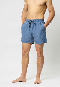 Mėlyni plaukimo šortai su raiščiu liemeniui, šoninėmis kišenėmis, lygiu paviršiumi ir mažu logotipu kairėje kojoje. Klasikinis pjūvis, kelių ilgio dizainas.