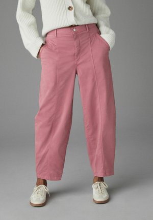 Personne portant un pantalon rose taille haute à jambes larges avec des coutures à l'avant, un pull en maille blanc et des baskets blanches à lacets, debout devant un fond gris.
