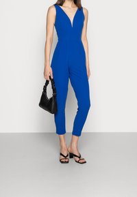 Mono azul con escote en V profundo y diseño ajustado, con pantalones de corte entallado. Combinado con un bolso negro y tacones de tiras negros.