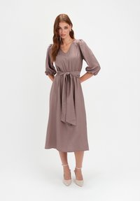 Kobieta ubrana w midi sukienkę w kolorze mauve z dekoltem w serek i bufkami, z przewiązanym paskiem w talii i beżowymi sandałami na kostkę, stojąca na białym tle.