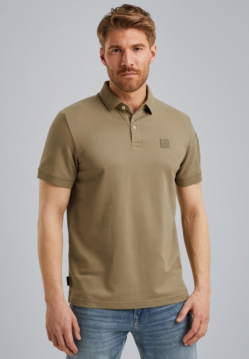 Khaki polo t-shirt lavet af bomuld, korte ærmer, to-knaps placket og et lille logo patch på venstre bryst. Bæret med blå jeans.