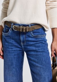 Ceinture en daim marron avec une boucle en argent, portée sur un jean en denim bleu foncé à coupe droite, avec des poches classiques et des détails de surpiqûres claires.