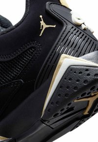 Svart sneaker med gulddetaljer, med en texturerad mesh-ovandel, perforerade sektioner och markerade detaljer på mellansulan, framhävda av en logotyp.