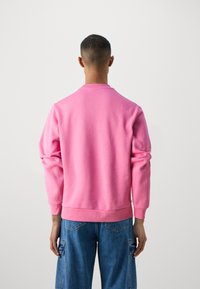 Sweat à capuche rose à manches longues, avec des poignets et un ourlet côtelés. Porté avec un pantalon cargo bleu comportant des poches latérales et de grandes poches au dos.