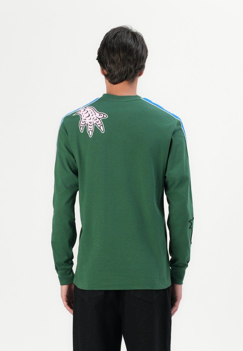 Zielona koszula z długim rękawem wykonana z bawełny. Posiada niebieskie paski na ramionach, różowy wzór inspirowany ośmiornicą z tyłu oraz czarne akcenty na rękawach.