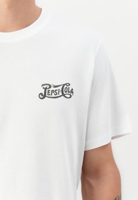 White T-shirt with vintage style black "Pepsi-Cola" logo on upper left chest, worn by a person with a visible forearm tattoo.