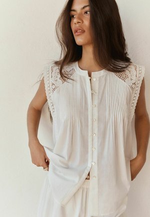 Vrouw in een mouwloze witte blouse met kanten schouders en knopen aan de voorkant, staand tegen een effen lichte muur, kijkend naar links.