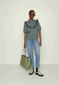 Γυναίκα με ανθισμένο μπλούζα με μαύρες λεπτομέρειες, ανοιχτό γαλάζιο τζιν, μπεζ μπότες, πράσινα γυαλιά ηλίου, κρατώντας μια μεγάλη ελαιόχρωμη τσάντα.