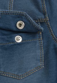 Détail de poche en denim avec une fermeture à pression argentée, des coutures dorées et un tissu bleu texturé. Présente une forme de poche triangulaire.