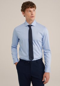 Világoskék, hosszú ujjú ing, galléros nyakkal, sötét navy nyakkendővel és sötét nadrággal párosítva. Az anyag simának tűnik, testhezálló fazonú.