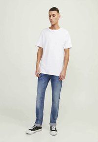 Ung man som står, iklädd en enkel vit t-shirt med kort ärm, blå uppvikta jeans och svarta sneakers med vita snörning på ljus bakgrund.