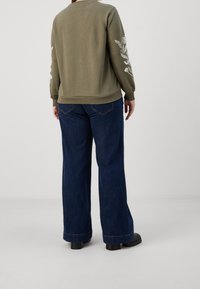 Felpa verde oliva con ricami floreali sulle maniche, abbinata a jeans a gamba larga in denim scuro e calzature nere chunky.