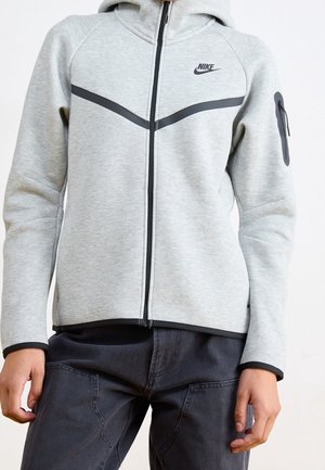 Jasnoszara bluza Nike na suwak z czarnymi detalami i kieszenią na suwak na lewym rękawie, noszona z ciemnymi spodniami.