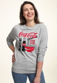 Felpa grigia con una grafica colorata della Coca-Cola, una ciotola di noodles e testo giapponese. Testo e immagini in rosso e nero.