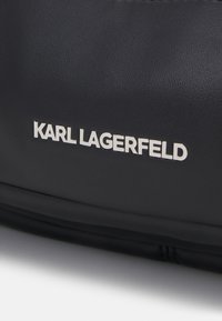 Melna ādas rokassoma ar teksturētu apdari. Priekšpusē ir izteikta balta "KARL LAGERFELD" logo. Vienkāršs, elegants dizains.