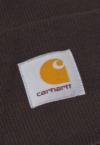 Ruskea neulottu kangas, jossa on neliömäinen valkoinen etiketti, jossa on keltainen logo ja "carhartt" tummanpunaisella tekstillä alareunassa.
