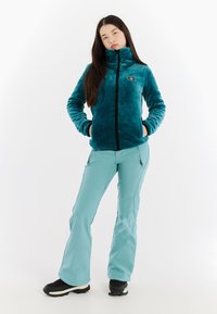 Teal fleece jack met hoge kraag en rits, gecombineerd met lichtblauwe waterdichte broek en zwarte laarzen. Gladde stof, slim fit ontwerp.