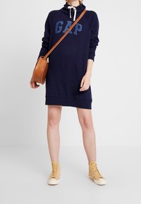 Granatowa sukienka dresowa z kapturem typu komin, z dżetami tworzącymi logo "GAP" w odcieniu niebieskiego brokatu, zestawiona z beżowymi trampkami i pasującą torebką na ramię.