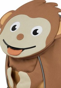 Mochila en forma de mono, hecha de tela marrón y beige; presenta una cara sonriente con ojos grandes, un detalle de lengua y orejas redondeadas. Cierre con cremallera.
