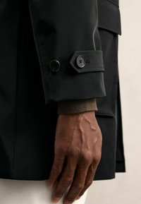 Fekete vízálló dzseki ujja gombos mandzsettával és a csuklórészből kilátszó kontrasztos kötött anyaggal; sima textúra és testre szabott kialakítás.