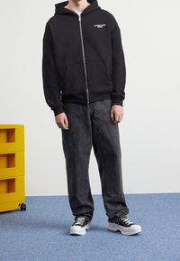 Black zip-up hoodie with front pockets and logo detail, paired with baggy dark gray pants and black-and-white sneakers on a blue floor.