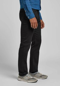 Jeans de mezclilla negra con corte ajustado, que cuentan con cinco bolsillos y una textura suave. Combinados con zapatos deportivos grises y una camisa de manga larga azul.