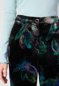Pantaloni din catifea neagră cu un imprimeu colorat de pene, cu închidere cu nasture și o suprafață moale, texturată. Bluză ușoară, albastră deschisă, cu mâneci lungi și manșete volan.