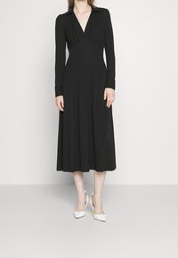 Robe noire à manches longues avec un décolleté en V, un corsage plissé et une jupe évasée. Portée avec des escarpins blancs à bout pointu dotés d'accents verts.