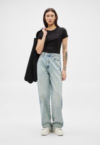 Ung kvinde med langt mørkt hår iført sort t-shirt, lyseblå vidde bukser, hvide sneakers, holder sort jakke over skulderen mod en ensfarvet baggrund.