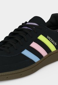 Baskets Adidas Spezial noires avec semelle marron et bandes pastel jaune, rose et bleue, avec lacets noirs et détail violet sur la languette.