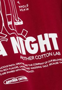 Punainen puuvillapaita, jossa on valkoista graafista tekstiä ja kuvituksia, mukaan lukien lauseet "YÖ" ja "kokojyvä", sekä logo alareunassa.