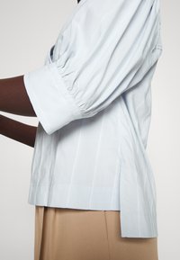 Lyseblå, stripete skjorte med puffede ermer, sidesliss ved kanten, og en glatt tekstur som står i kontrast til beige bukser.