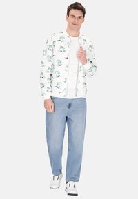 Hvid zip-up jakke med tropisk palme mønster, båret over en hvid T-shirt, parret med lyseblå jeans og hvide sneakers.