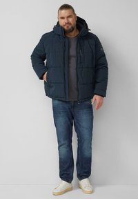 Granatowa puchowa kurtka z kapturem, z pikowanymi panelami i kieszenią na suwak na piersi. Połączona z szarą koszulą i niebieskimi dżinsami.