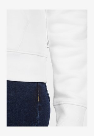 Hvid sweatshirt med ribbede manchetter, der har et lille mærkat på siden. Båret sammen med mørkeblå denim jeans.