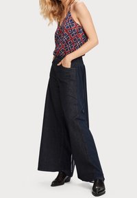 Haut sans manches à motifs en rouge, bleu marine et blanc ; pantalon large bleu marine avec plis sur les côtés ; modèle portant des chaussures foncées, les mains dans les poches.