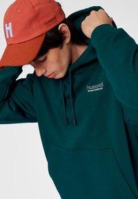 Dunkelgrüner Hoodie mit Fronttasche und Logo. Orangefarbene Kappe mit weißem "H." Der Hoodie verfügt über Kordelzüge und gerippte Bündchen.
