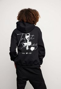 Felpa nera con cappuccio e stampa grafica sul retro che riporta la parola "Trust" e presenta illustrazioni, abbinata a pantaloni neri coordinati.