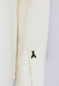 Camicia in lino di colore avorio con un evidente emblema di insetto nero sul polsino. Tessuto testurizzato, dettaglio della manica su misura.