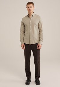 Helle beige Knopfbundbluse mit spitzem Kragen, glatter Textur, kombiniert mit dunkelbraunen maßgeschneiderten Hosen und schwarzen Schuhen. Minimale Markenkennzeichnung sichtbar.