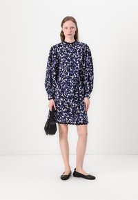 Blumenmusterkleid in Marineblau, Weiß und Helllila; mit Puffärmeln, tailliertem Bund und gewelltem Saum. Schwarze Handtasche und Ballerinas.