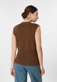 Γυναίκα με τα μαλλιά της σε κότσο, φορώντας καφέ αμάνικο τοπ και μπλε τζιν, φαίνεται από πίσω μπροστά σε λευκό απλό φόντο.